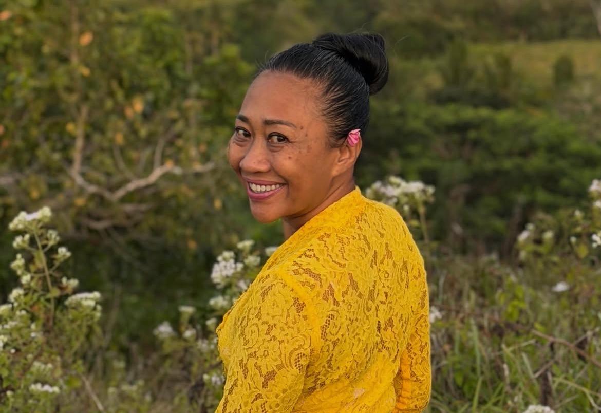 Smiling woman with dark hair in a bun wearing a yellow lace top standing outdoors among wildflowers and greenery